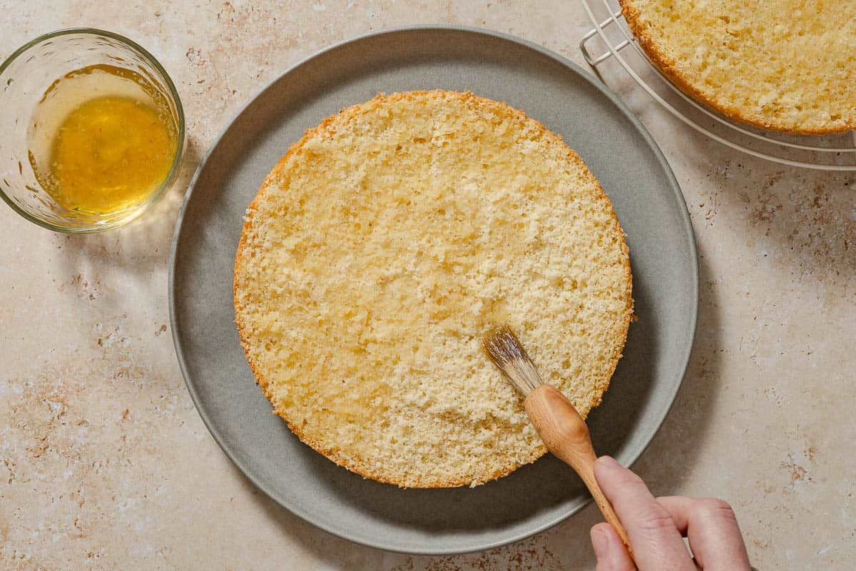 An overhead photo of the syrup being brushed onto the bottom half of the cassata cake on a platter. Next to this is a cup of the syrup, and the top half of the cake on a cooling rack.