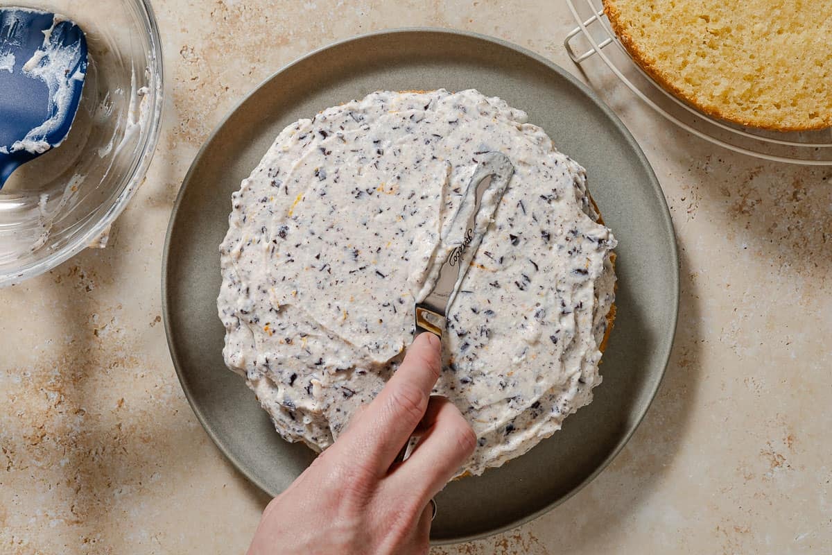 The filling for the cassata cake being spread on the bottom half of the cake with a knife. Next to this is the empty filling bowl with a spatula, and the top half of the cake on a wire rack.