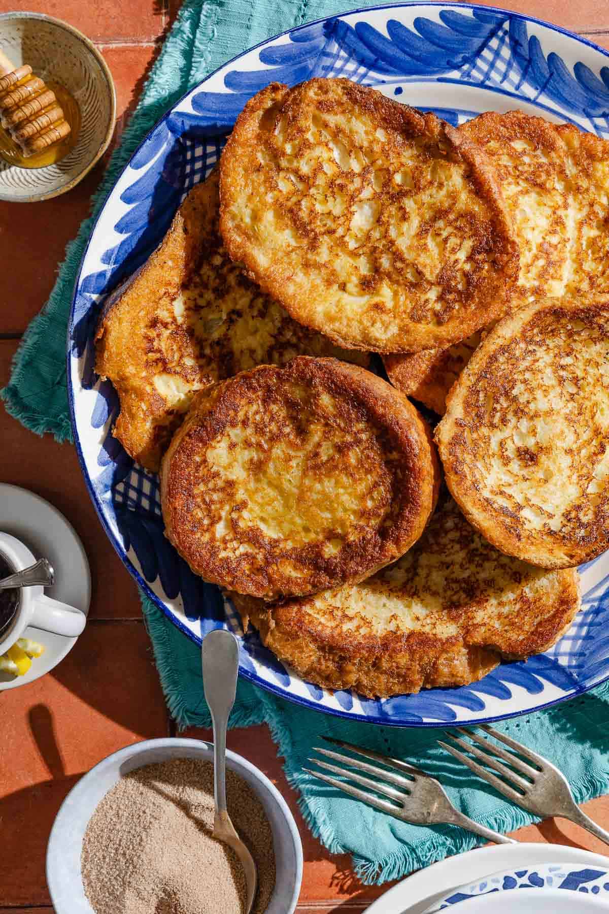 Several torrijas on a serving platter surrounded by bowls of honey and cinnamon sugar, a cup of coffee, a stack of plates and 2 forks.