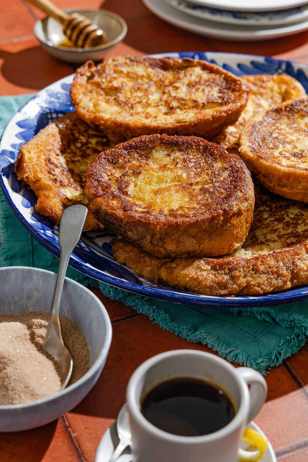 Several torrijas on a serving platter surrounded by bowls of honey and cinnamon sugar, and a cup of coffee.