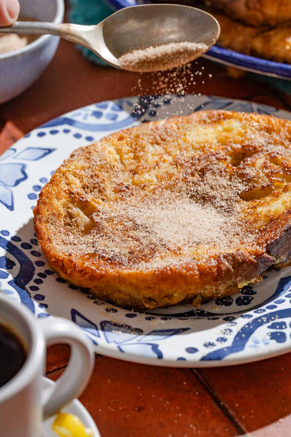 Cinnamon sugar being sprinkled from a spoon onto a torrija on a plate.