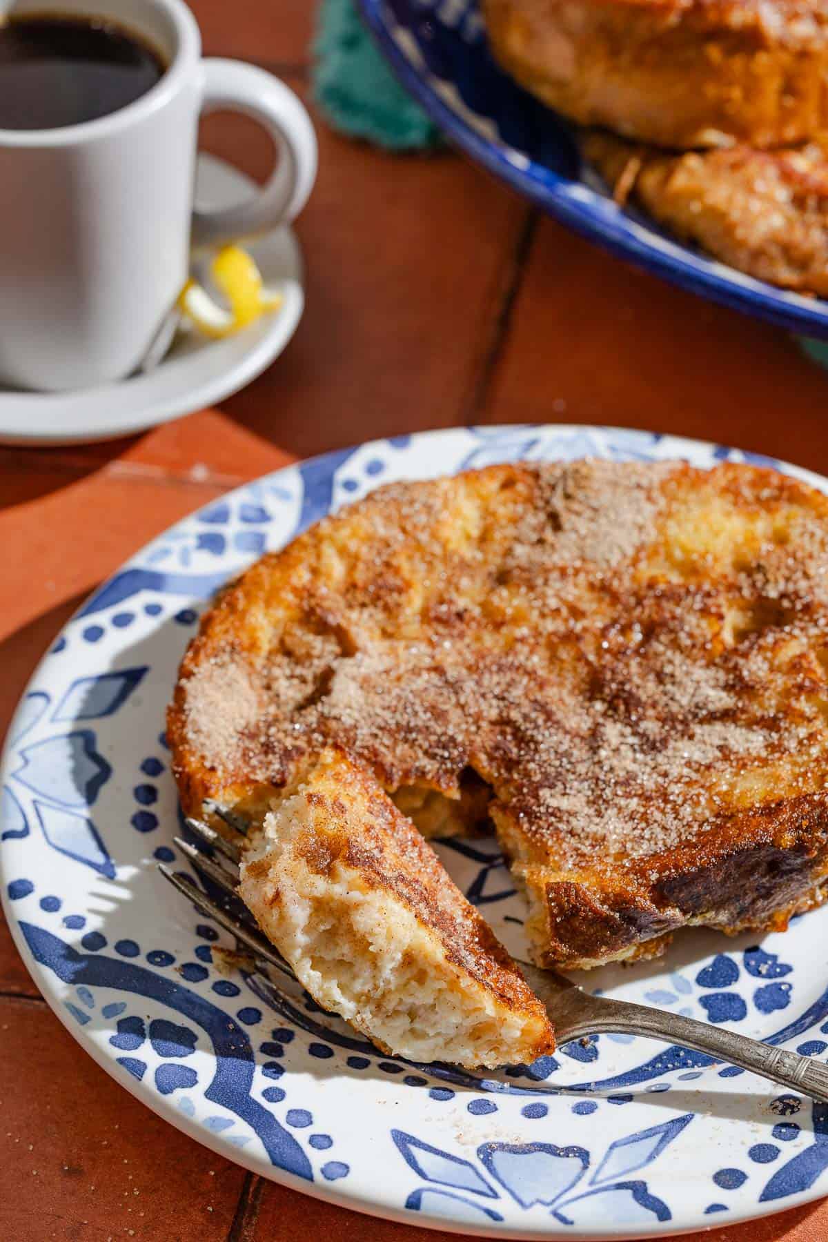 A torrija on a plate with a piece taken out of it with a fork. Next to this is a serving platter with the rest of the torrijas, and a cup of coffee.