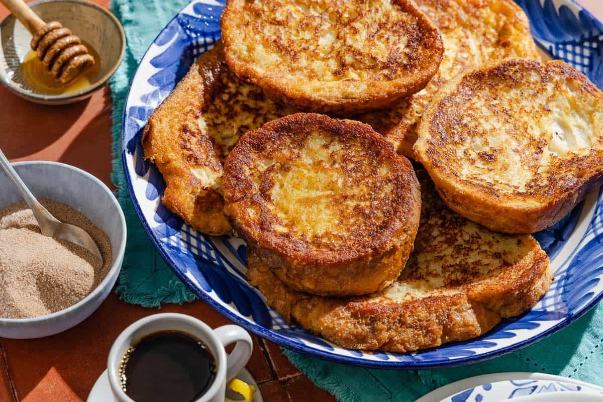 A close up photo of several torrijas on a serving platter surrounded by bowls of honey and cinnamon sugar, and a cup of coffee.
