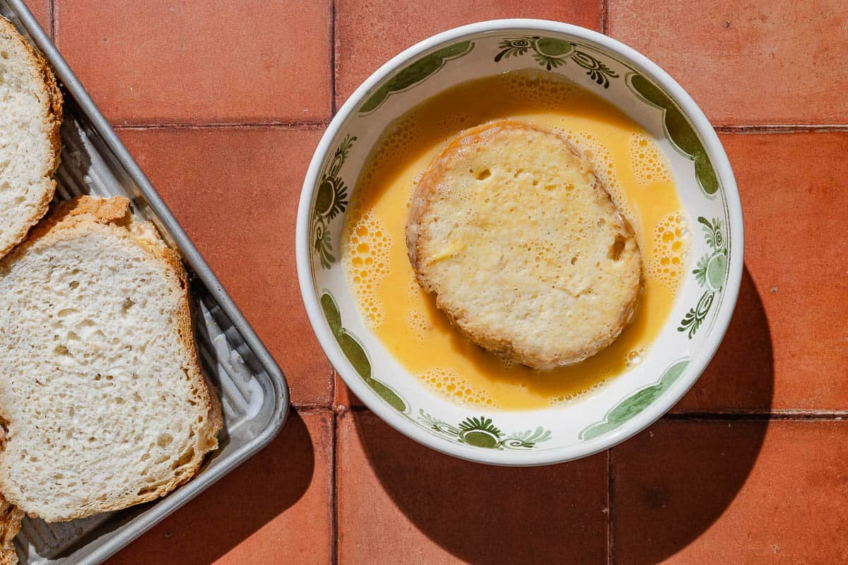 A slice of bread being dipped in a bowl of scrambled eggs next to a baking sheet with more slices of bread.