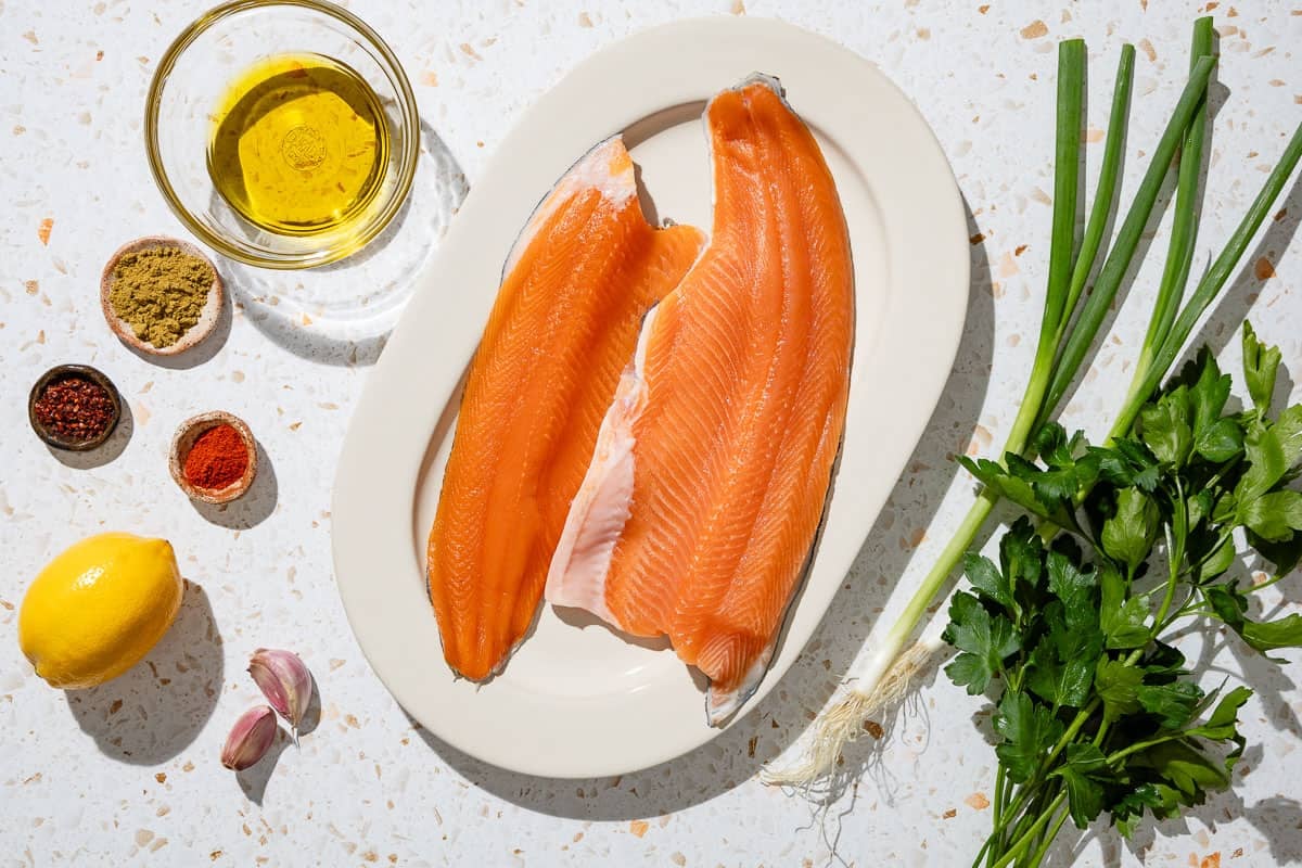 Ingredients for baked trout including trout fillets, olive oil, lemon, cumin, paprika, urfa biber, garlic, green onions, parsley, salt and pepper.