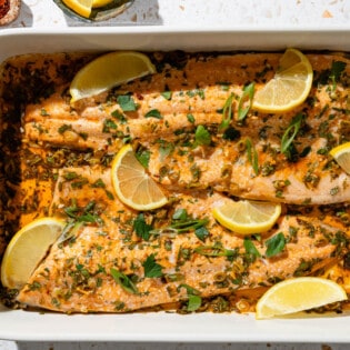 An overhead photo of baked trout garnished with parsley and green onions in a baking dish. This is surrounded by small bowls of green onions, urfa biber pepper and lemon wedges.