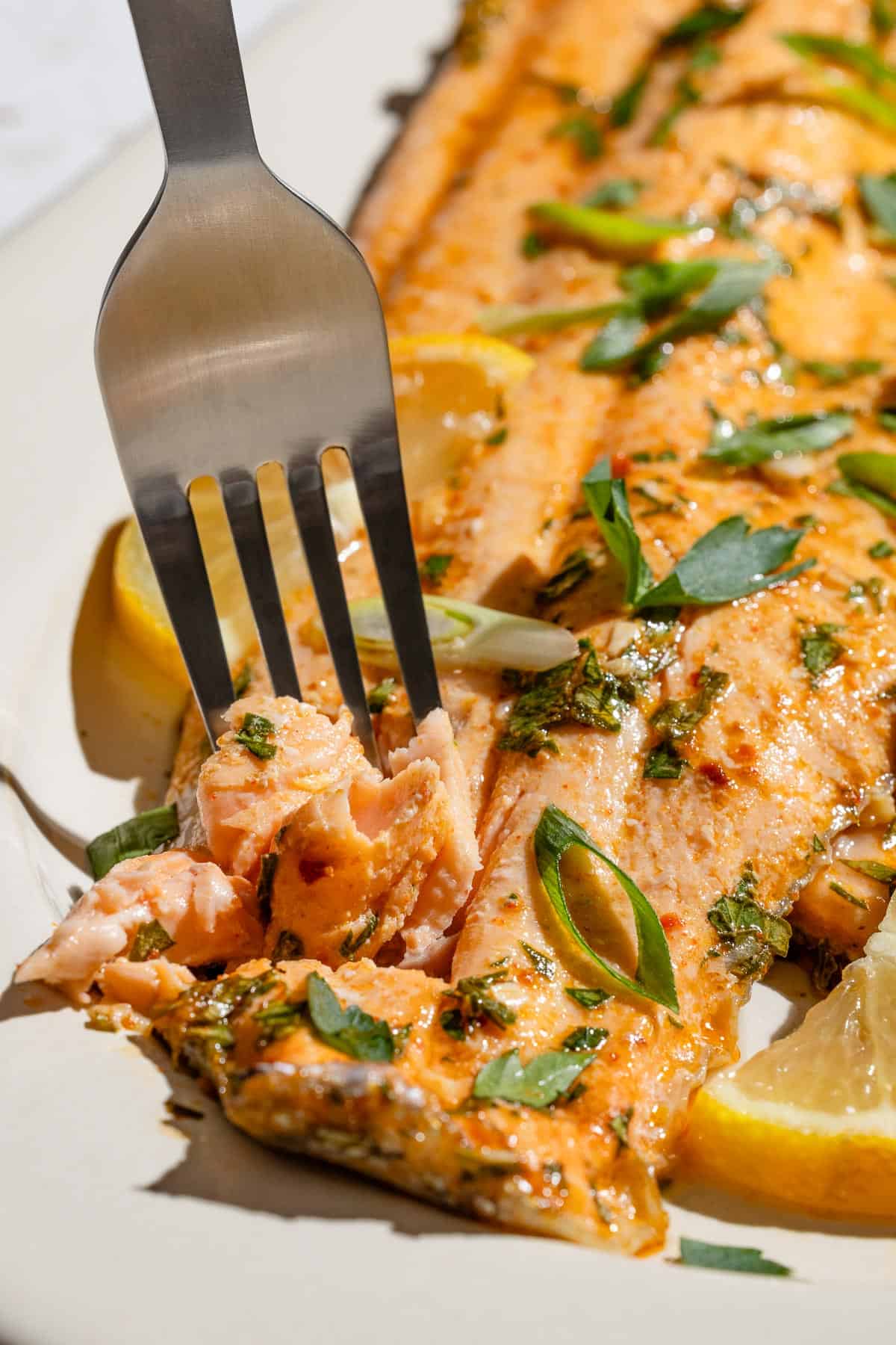 A close up of a fork digging into a baked trout fillet garnished with parsley, green onions and a lemon wedge.