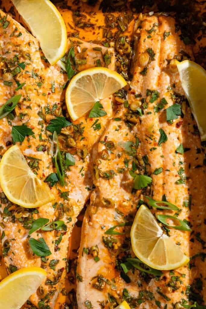 A close up of baked trout garnished with parsley, green onions and lemon wedges.