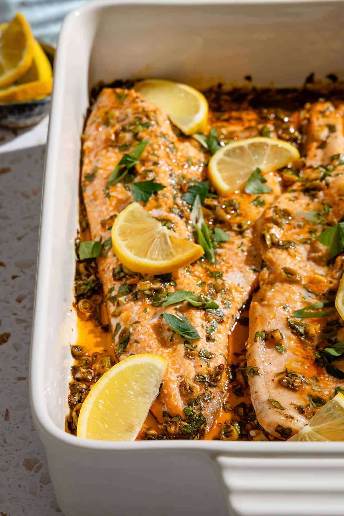 A close up of baked trout garnished with parsley and green onions in a baking dish next to a small bowl of lemon wedges.