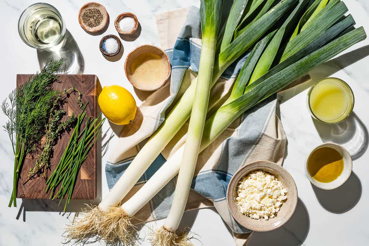 Ingredients for braised leeks including leeks, olive oil, sugar, salt, pepper, lemon, white wine, vegetable broth, fresh thyme, fresh chives, fresh dill, and feta cheese.