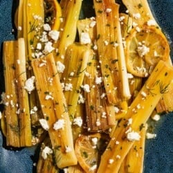 An overhead photo of braised leeks garnished with dill, crumbled feta and lemon slices on a blue serving platter.