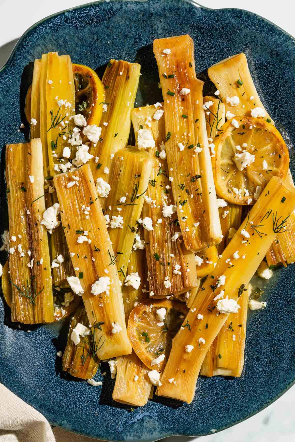 An overhead photo of braised leeks garnished with dill, crumbled feta and lemon slices on a blue serving platter.