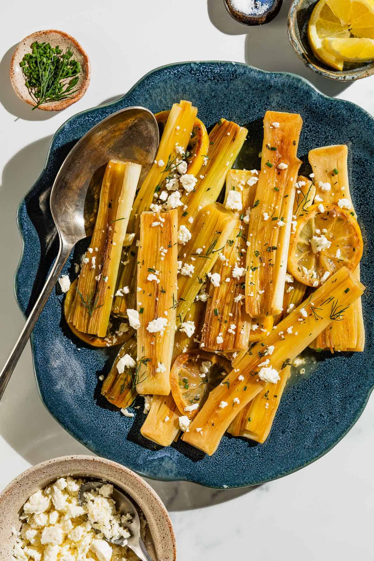 An overhead photo of braised leeks garnished with dill, crumbled feta and lemon slices on a blue serving platter with a serving spoon. This is surrounded by small bowls of dill and chives, salt, lemon wedges and crumbled feta cheese.