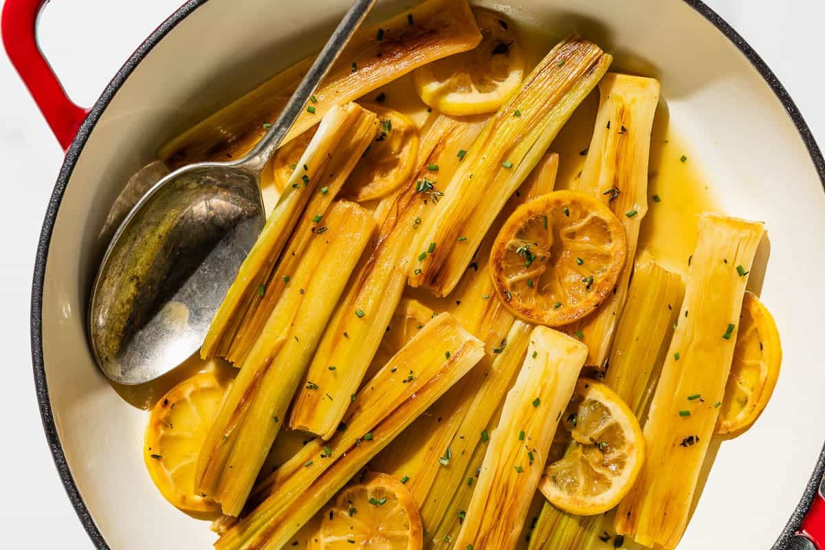 A close up of braised leeks topped with lemon slices and chives in a skillet with a serving spoon.