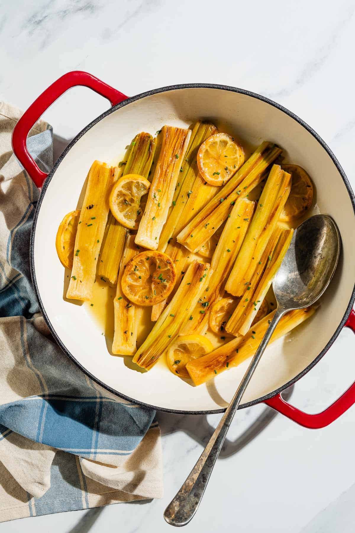 An overhead photo of raised leeks topped with lemon slices and chives in a skillet with a serving spoon. Next to this is a cloth napkin.