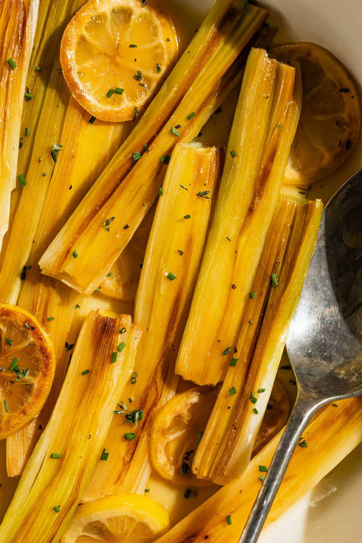 A close up photo of Braised leeks topped with lemon slices and chives in a skillet with a serving spoon.