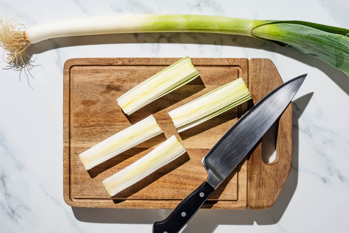 A leek cut into 4 pieces on a cutting board with a knife. Next to this is an entire leek.