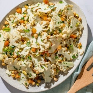An overhead photo of a shaved cauliflower salad in a serving bowl. Next to this is a set of wooden serving utensils on a cloth napkin.