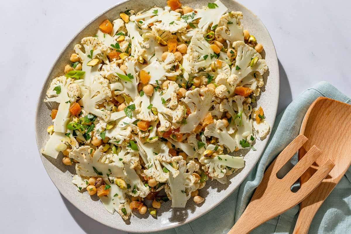 An overhead photo of a shaved cauliflower salad in a serving bowl. Next to this is a set of wooden serving utensils on a cloth napkin.