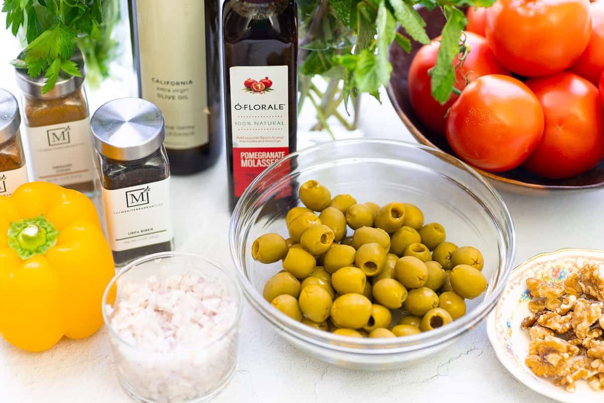 Ingredients for olive salad including castelvetrano olives, pomegranate molasses, urfa biber, paprika, cumin, olive oil, shallots, tomato, a bell pepper, parsley, mint, and walnuts.