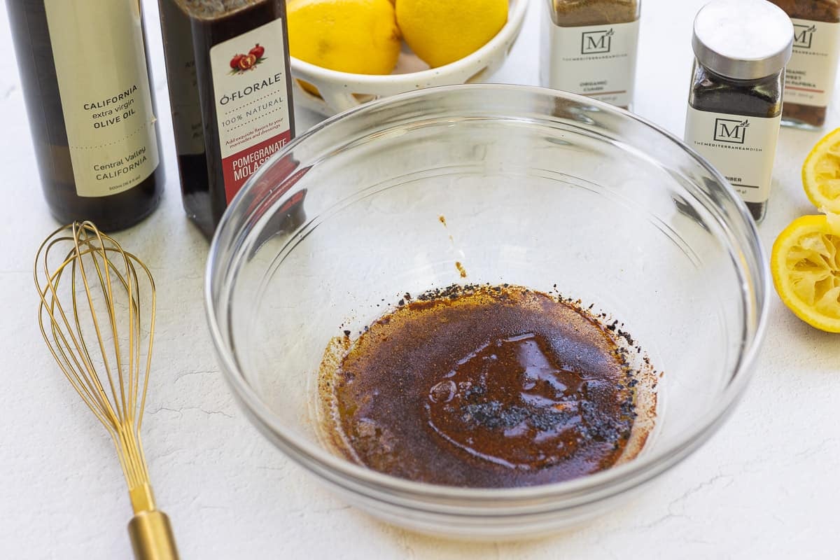 The dressing for the olive salad in a mixing bowl. Next to this is a whisk, 2 juiced lemon halves, bottles of olive oil and pomegranate molasses, jars of paprika, cumin and urfa biber, and an bowl of lemons.