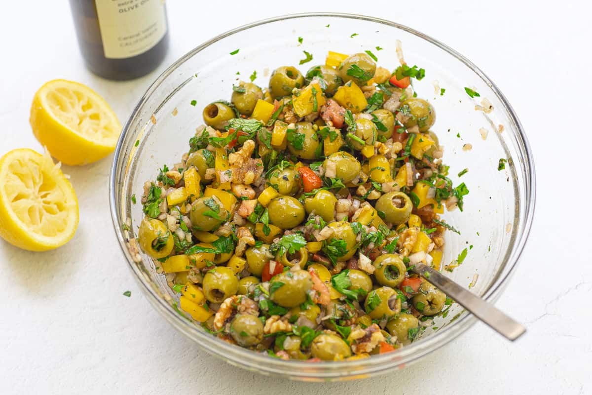 A close up of olive salad in a mixing bowl with a spoon. Next to this is a bottle of olive oil and 2 juiced lemon halves.