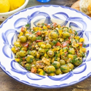 A close up of olive salad in a serving bowl. Next to this is a bowl of lemons, some crusty bread and three forks.