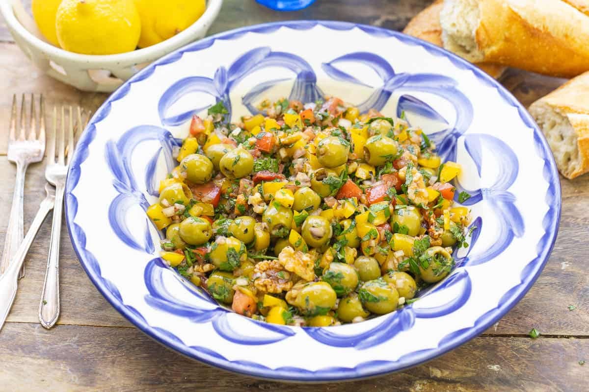 A close up of olive salad in a serving bowl. Next to this is a bowl of lemons, some crusty bread and three forks.