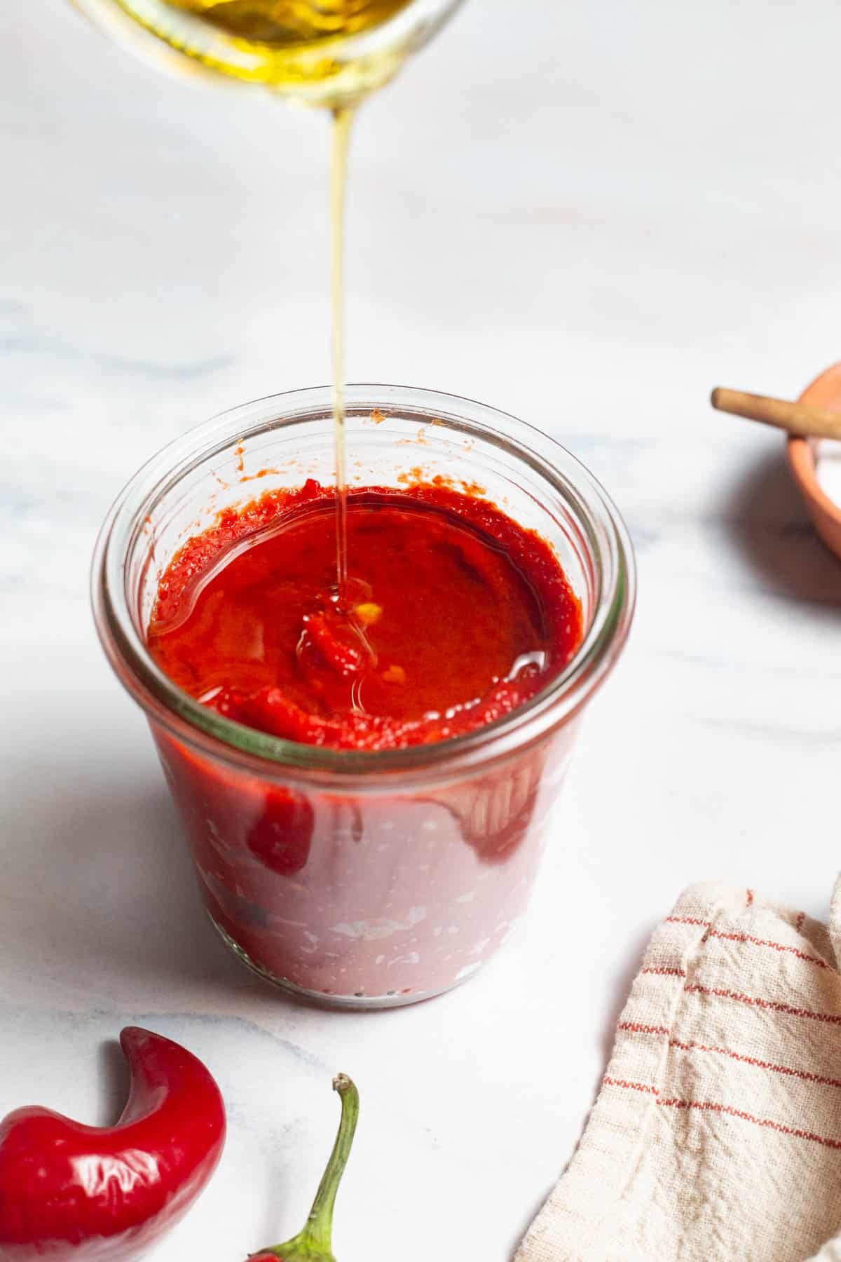 Olive oil being poured in to a jar of red pepper past. Next to this is a kitchen towel, bowl of salt and 2 red chili peppers.