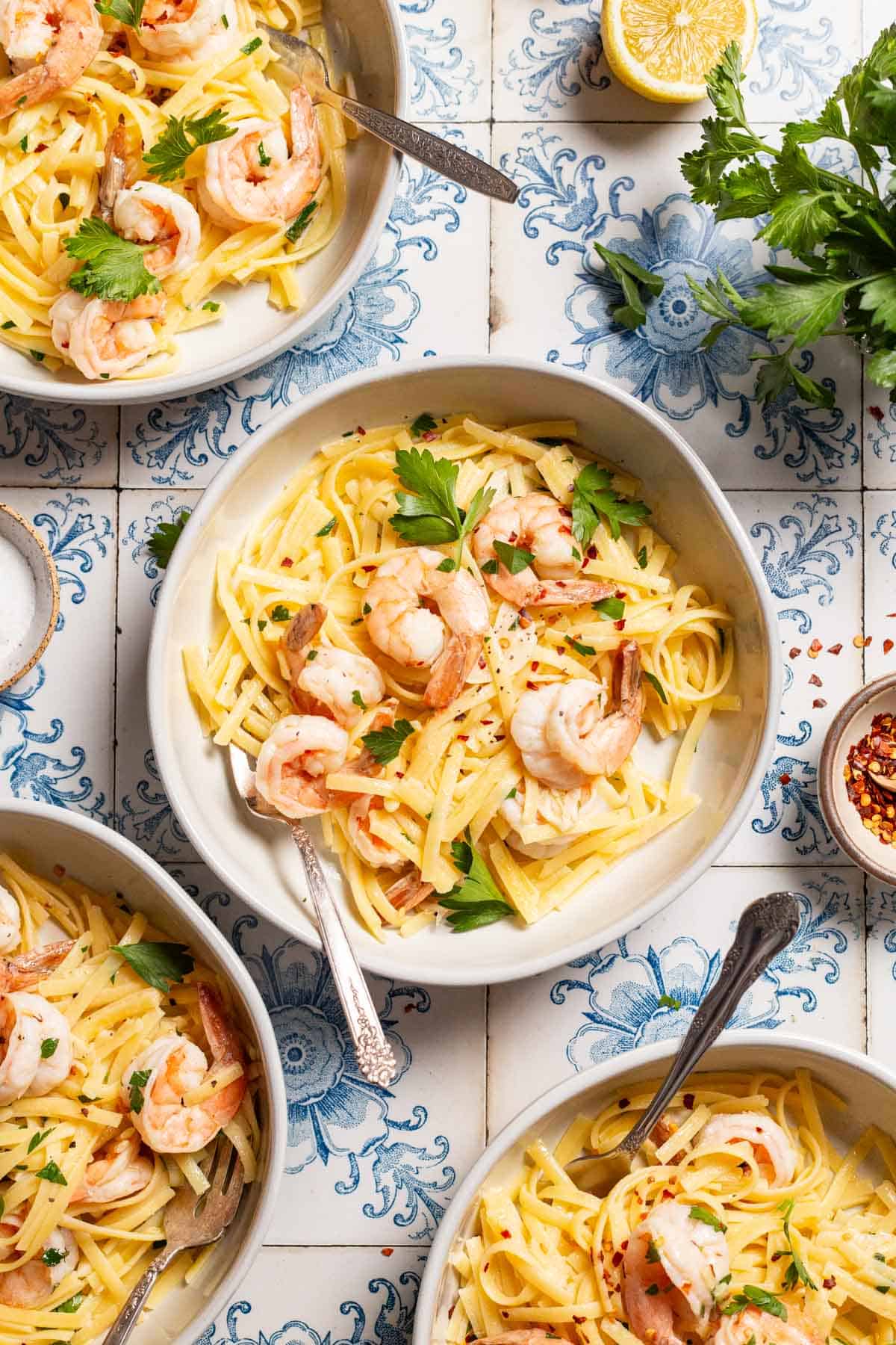 An overhead photo of four bowls of shrimp linguine with forks. Next to this are small bowls of salt and red pepper flakes, a lemon half, and parsley.