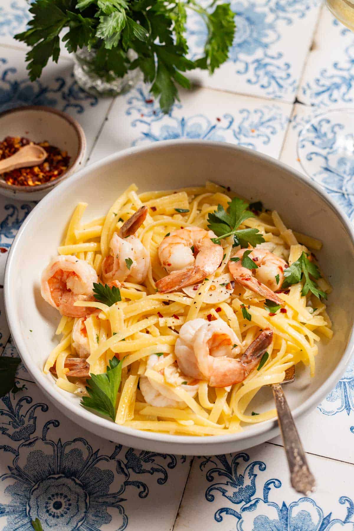 Shrimp linguine in a bowl with a fork next to a bowl of red pepper flakes and parsley.