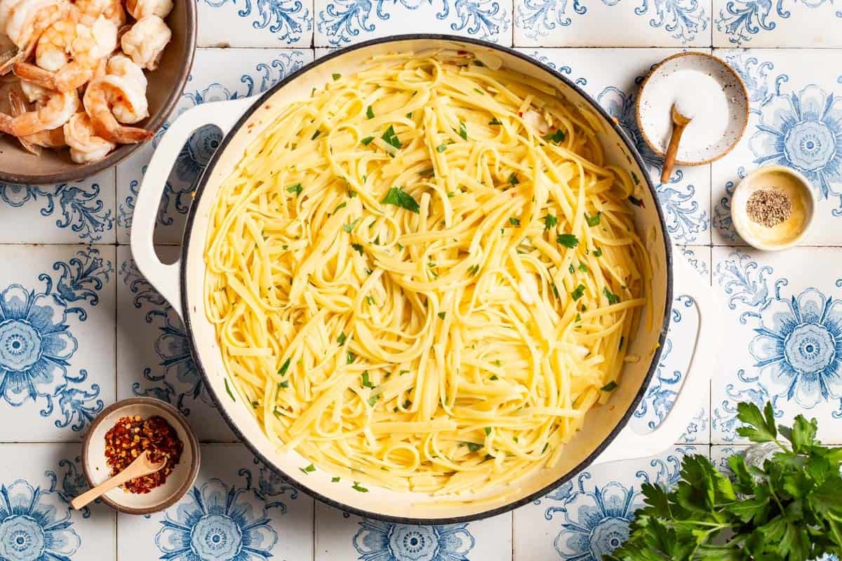 An overhead photo of linguine in sauce in a large pot. Surrounding this are bowls of shrimp, salt, pepper and red pepper flakes and parsley.