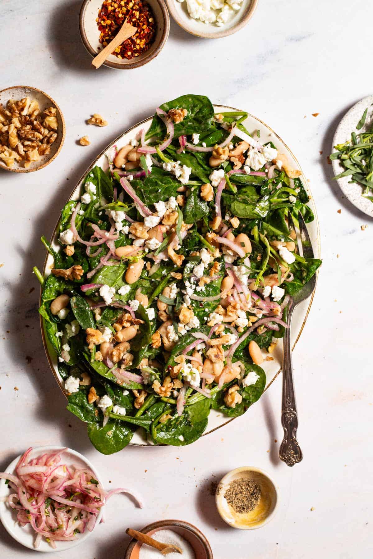 An overhead photo of the wilted spinach salad on a serving platter with a fork. Surrounding this are small bowls of salt, pepper, walnuts, pickled onions, tarragon, goat cheese, and red pepper flakes.