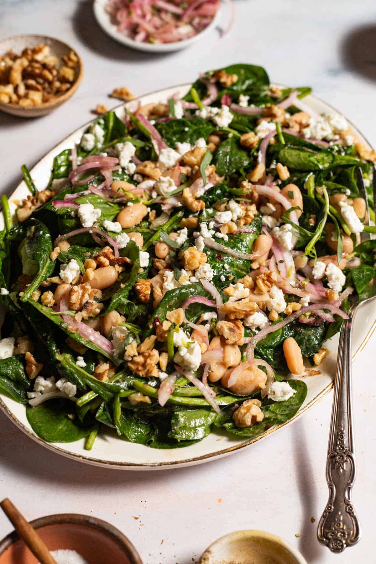 The wilted spinach salad on a serving platter with a fork. Surrounding this are small bowls of salt, walnuts, and pickled onions.