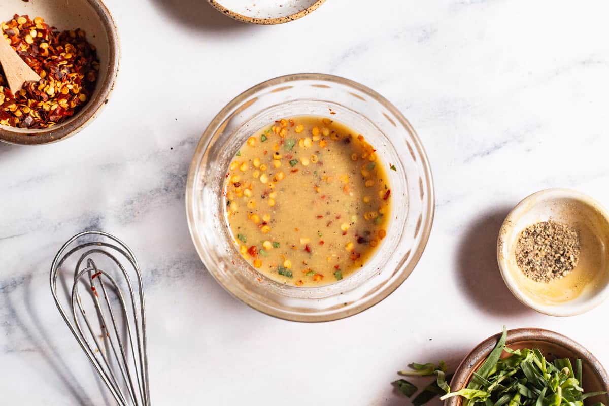 The dressing for the wilted spinach salad in a bowl next to a whisk and bowls of red pepper flakes, black pepper and tarragon.