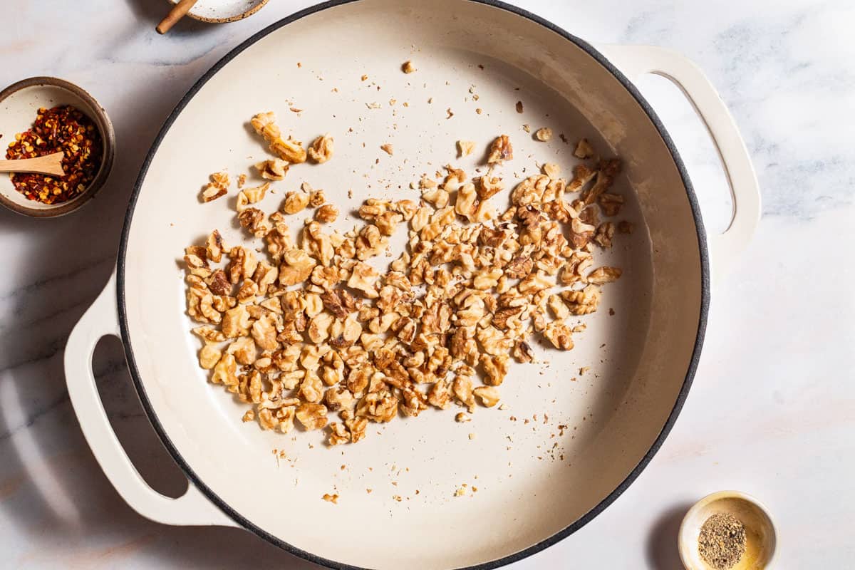 Walnuts being toasted in a skillet next to bowls of red pepper flakes and black pepper.