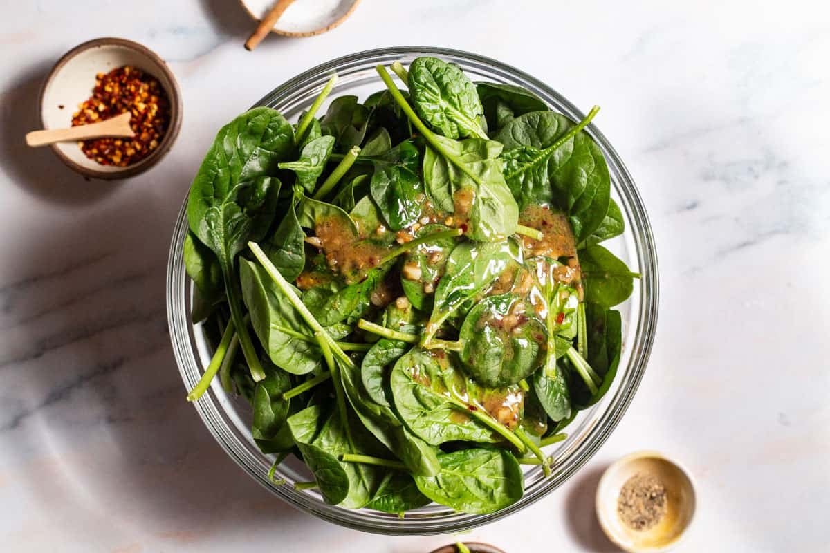 Spinach in a large bowl topped with the warmed dressing. Next to this are small bowls of red pepper flakes, salt and black pepper.