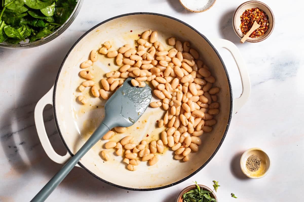 Cannellini bean being warmed in a skillet with a spatula. Surrounding this are bowls of spinach, salt, red pepper flakes, pepper, and tarragon.