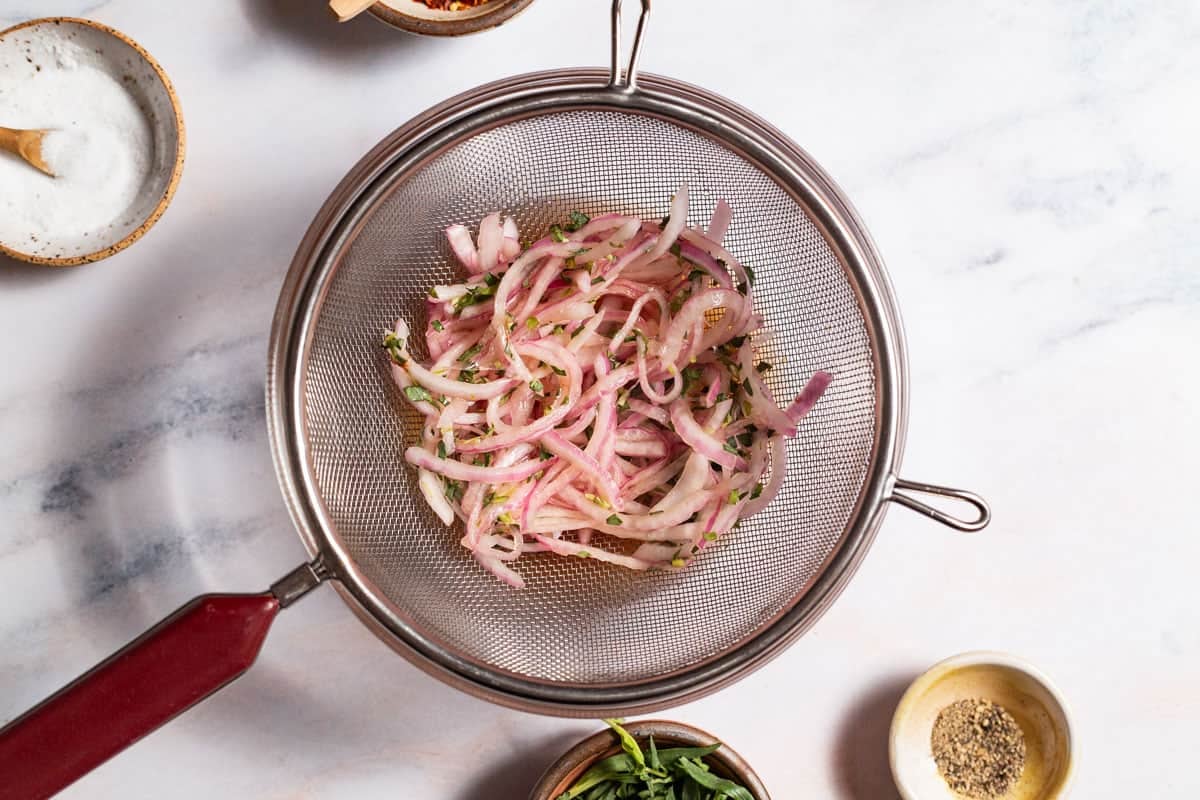 Pickled red onions in a colander next to small bowls of salt, pepper and tarragon.