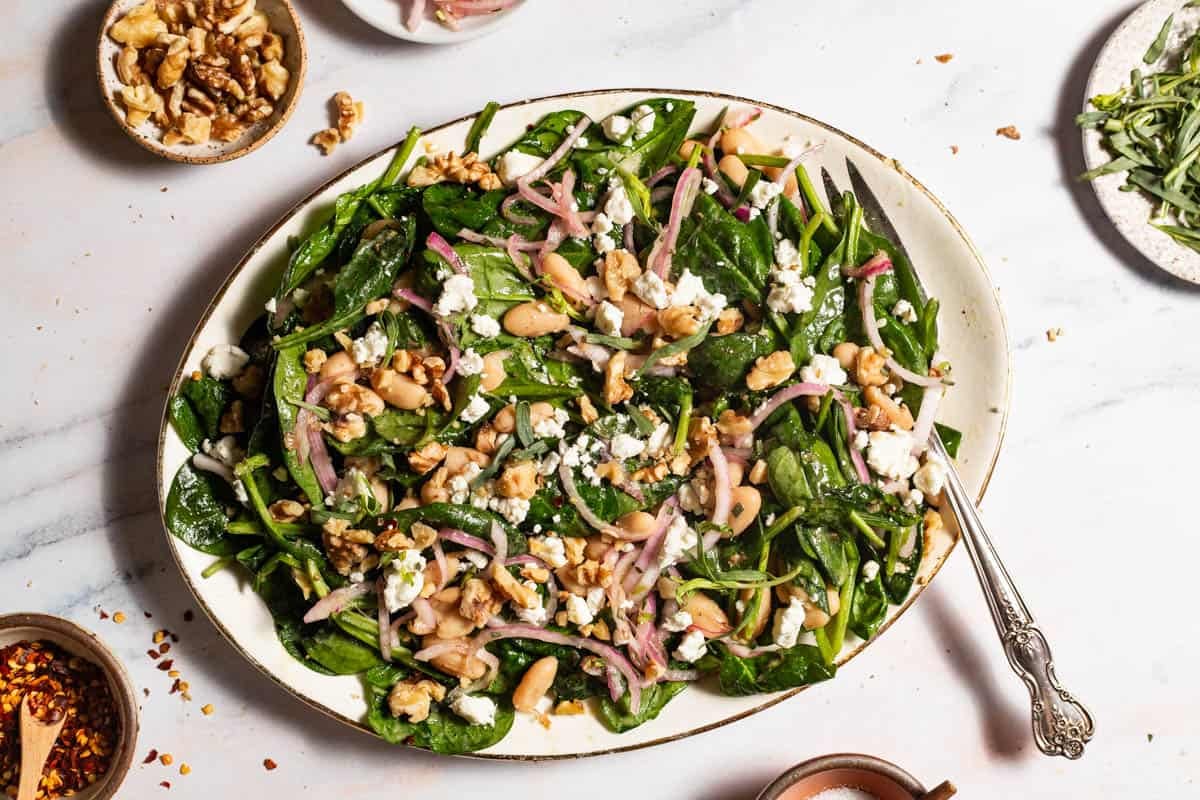 An overhead photo of the wilted spinach salad on a serving platter with a fork. Surrounding this are small bowls of walnuts, tarragon, and red pepper flakes.