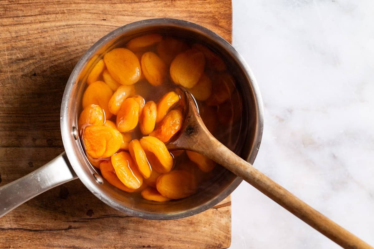 Dried apricots being poached in the syrup in a saucepan with a wooden spoon.