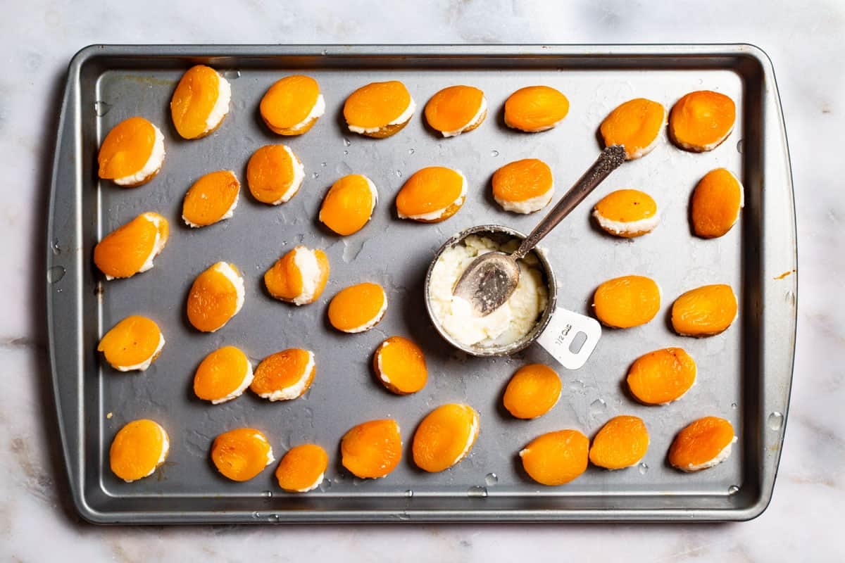 Dried apricots stuffed with ricotta on a baking sheet. Also on the baking sheet is a measuring cup with more ricotta and a spoon.