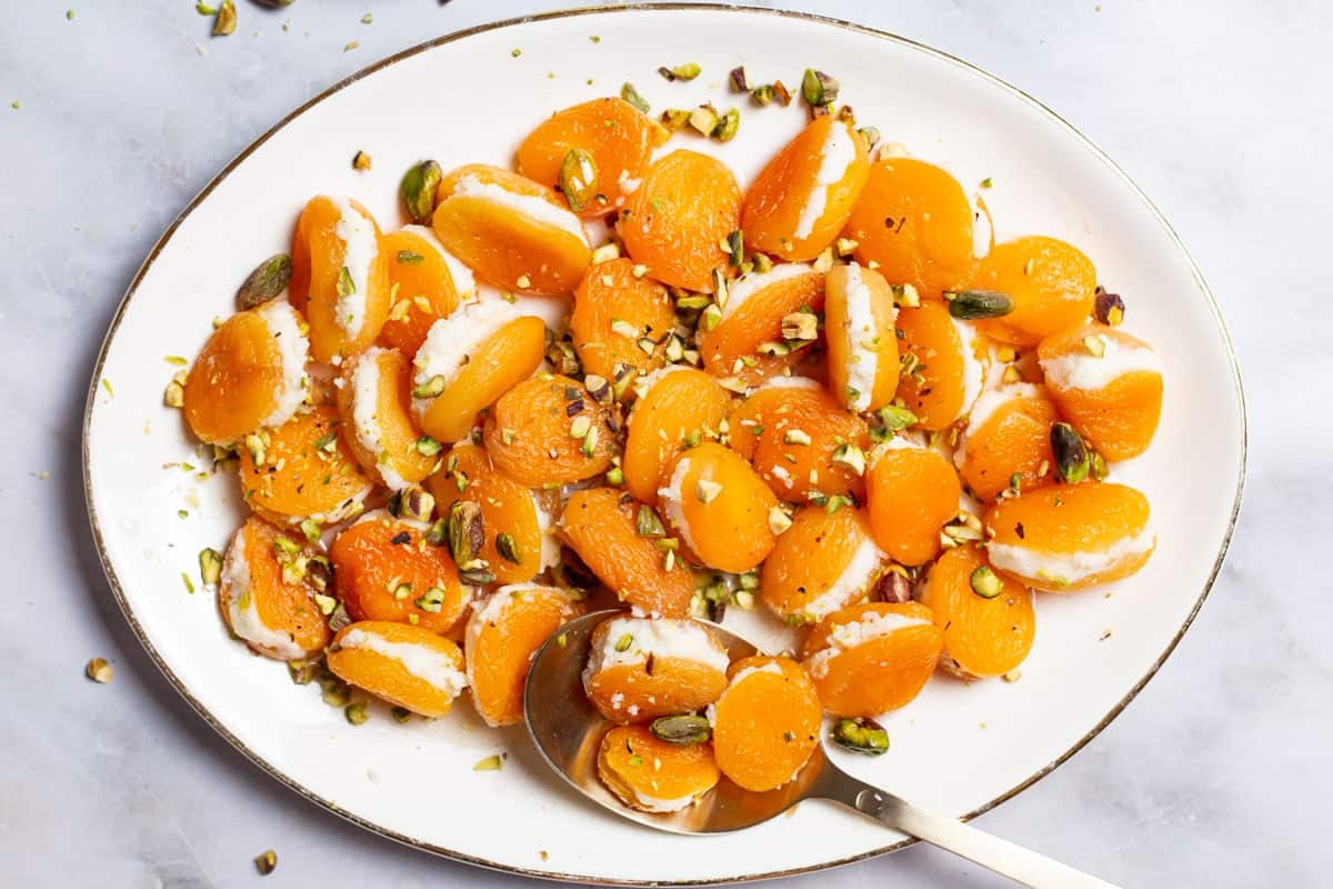 An overhead photo of stuffed apricots topped with pistachios on a serving platter with a spoon.
