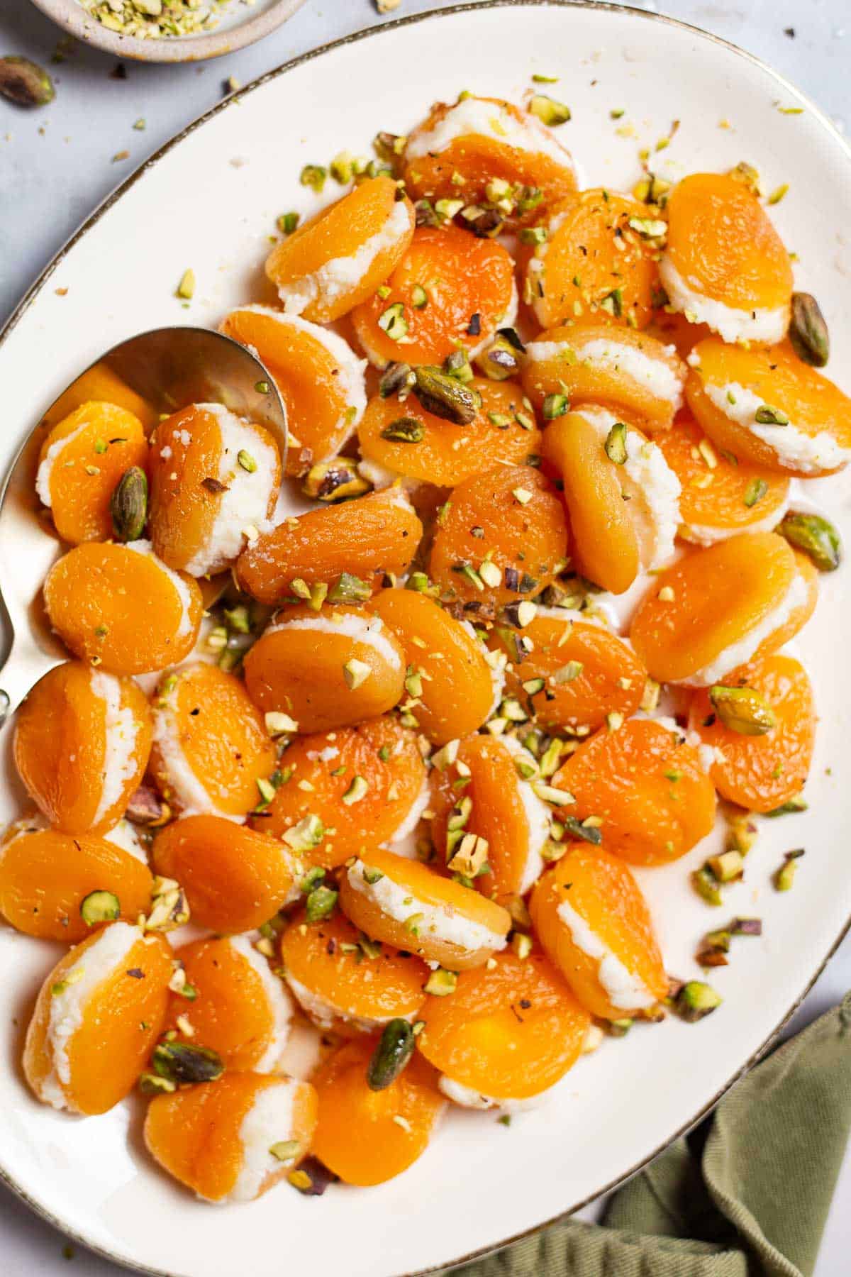 An overhead close up photo of stuffed apricots topped with pistachios on a serving platter with a spoon.