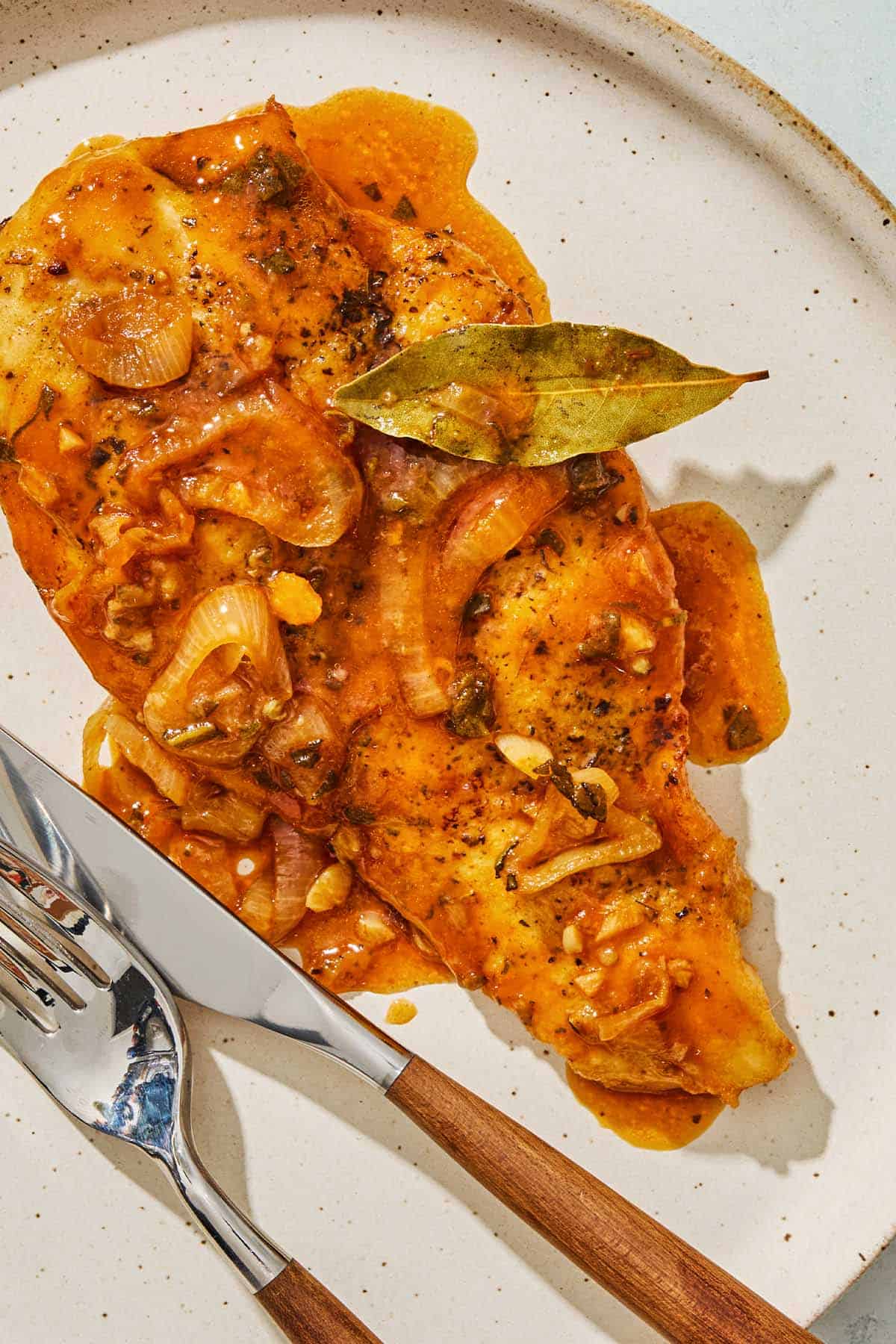 A close up photo of a braised chicken breast on a plate with a fork and knife.