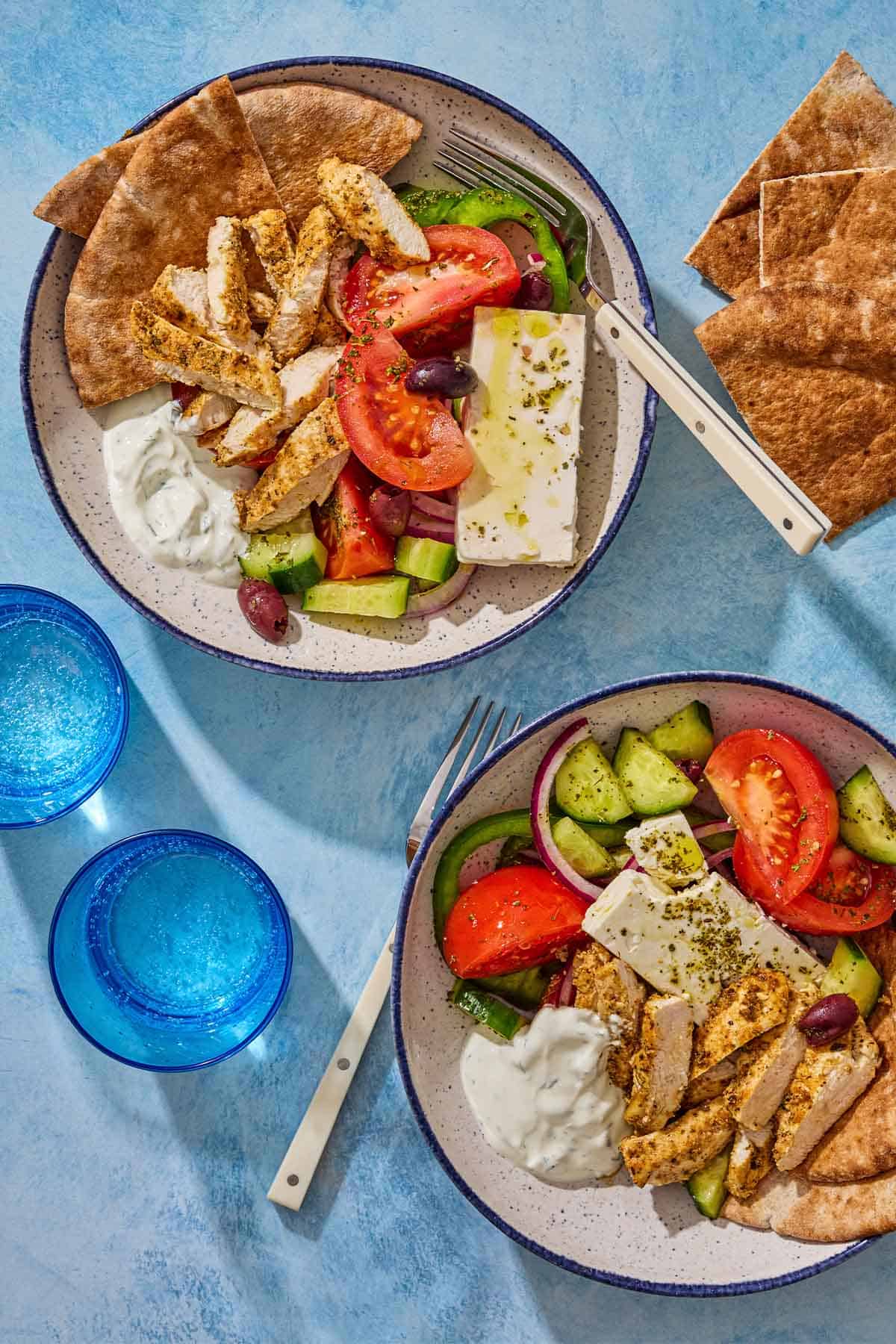 An overhead photo of 2 gyro bowls with forks. Next to these are 2 glasses of water and pieces of pita bread.
