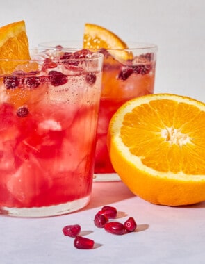 A close up of 2 pomegranate fizz mocktails garnished with pomegranate seeds and orange wedges next to more pomegranate seeds and 2 orange halves.