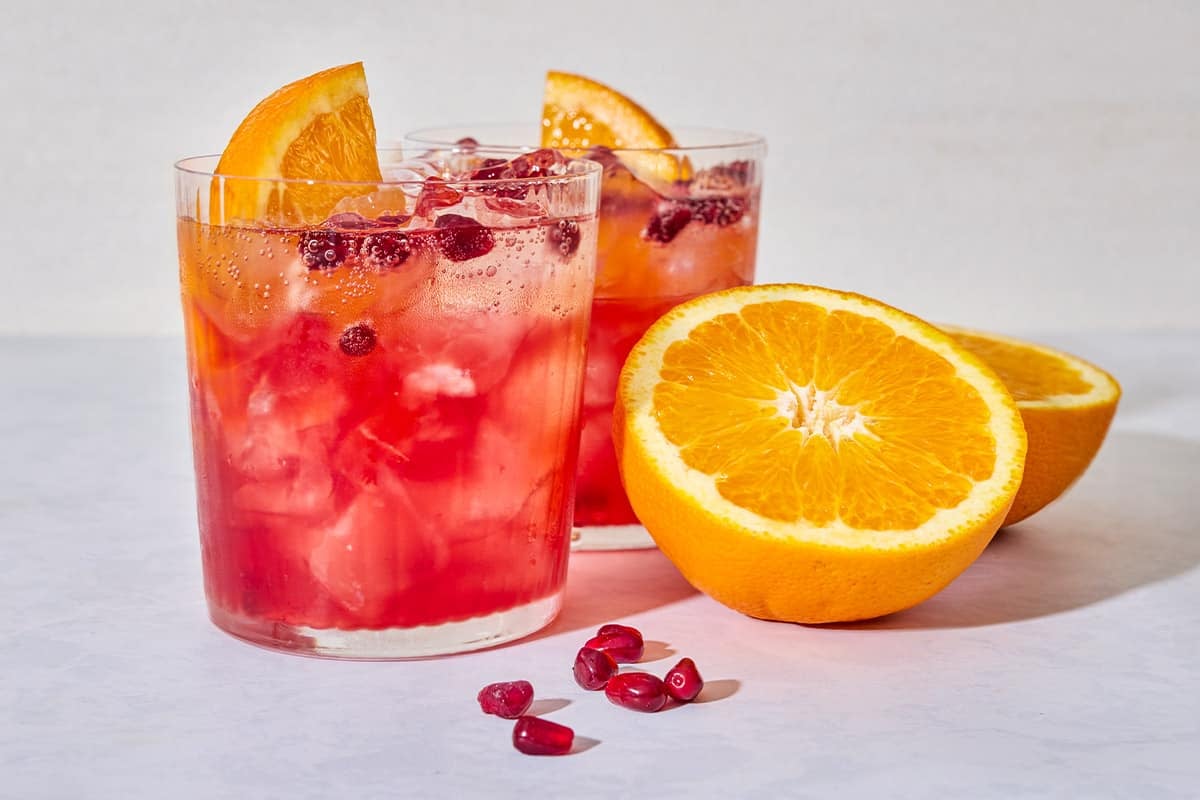 A close up of 2 pomegranate fizz mocktails garnished with pomegranate seeds and orange wedges next to more pomegranate seeds and 2 orange halves.