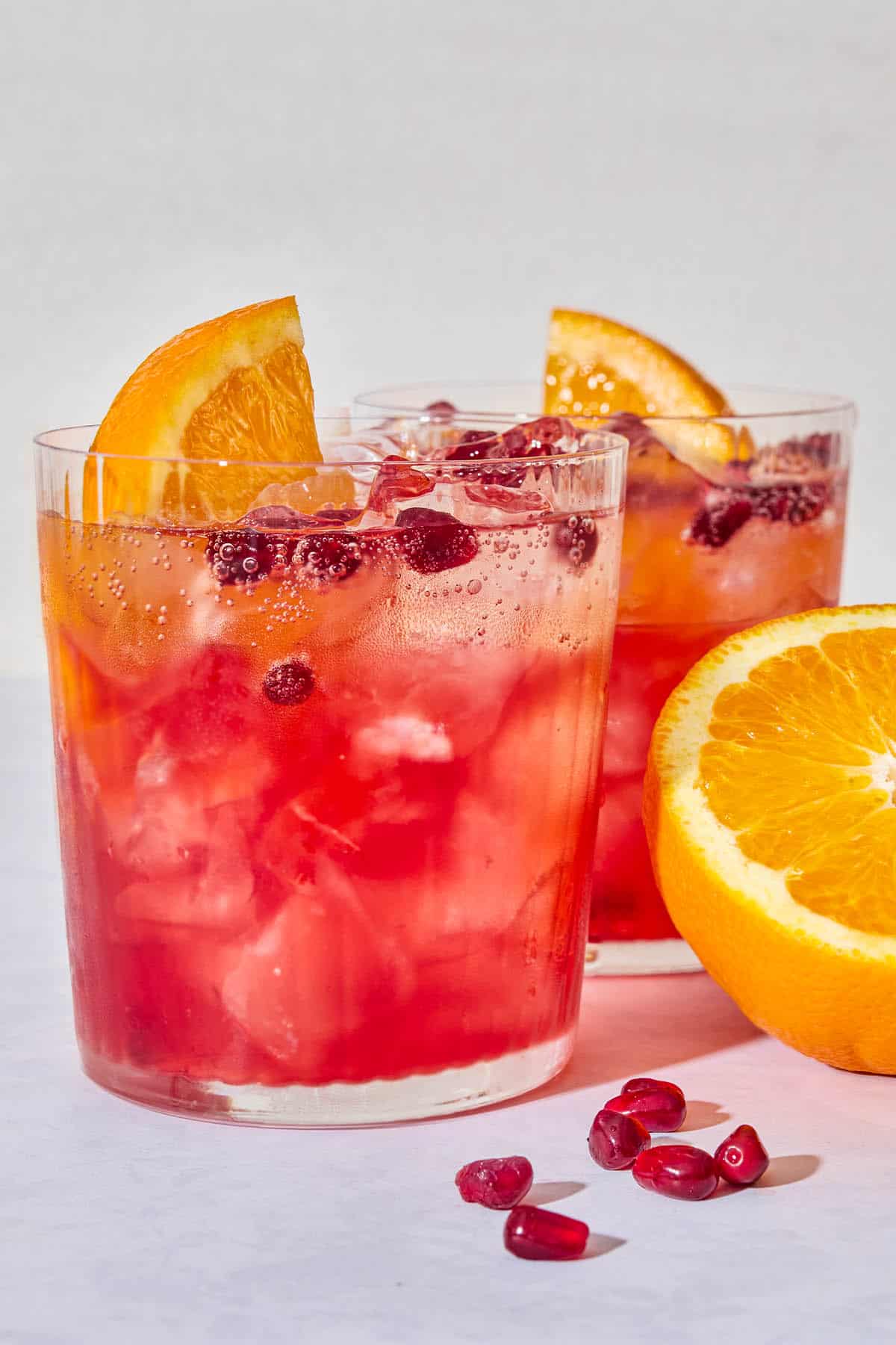 A close up of 2 pomegranate fizz mocktails garnished with pomegranate seeds and orange wedges next to more pomegranate seeds and a half of an orange.