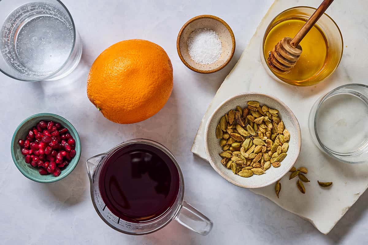 Ingredients for the pomegranate mocktail recipe including pomegranate juice, club soda, pomegranate seeds, an orange, green cardamom pods, honey, water, and kosher salt.